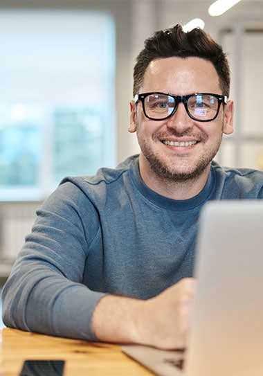 Happy man working on a laptop in an office setting, smiling at the camera.